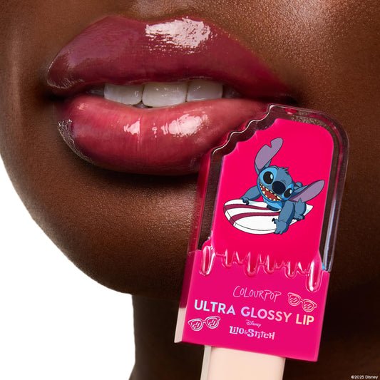 Surfer Vibes Ultra Glossy Lip by ColourPop glazing lips with a high-shine rosy‑pink finish in a tight close-up; bright pink tube with Disney Lilo & Stitch’s Stitch graphic is held by the mouth, showcasing the smooth, cushiony gloss.