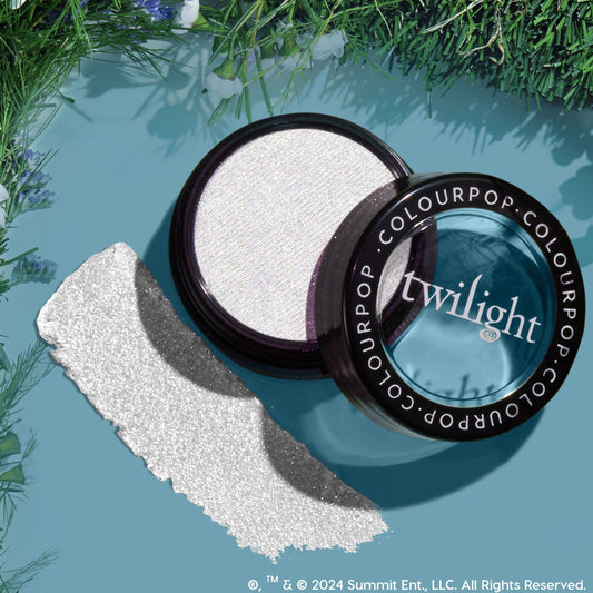 Vampire Skin ColourPop Super Shock Highlighter gleams in an open jar, showing an icy white, sparkly pan with the Twilight-branded lid and a bold swatch, styled as a flat lay on a teal backdrop bordered with green foliage.