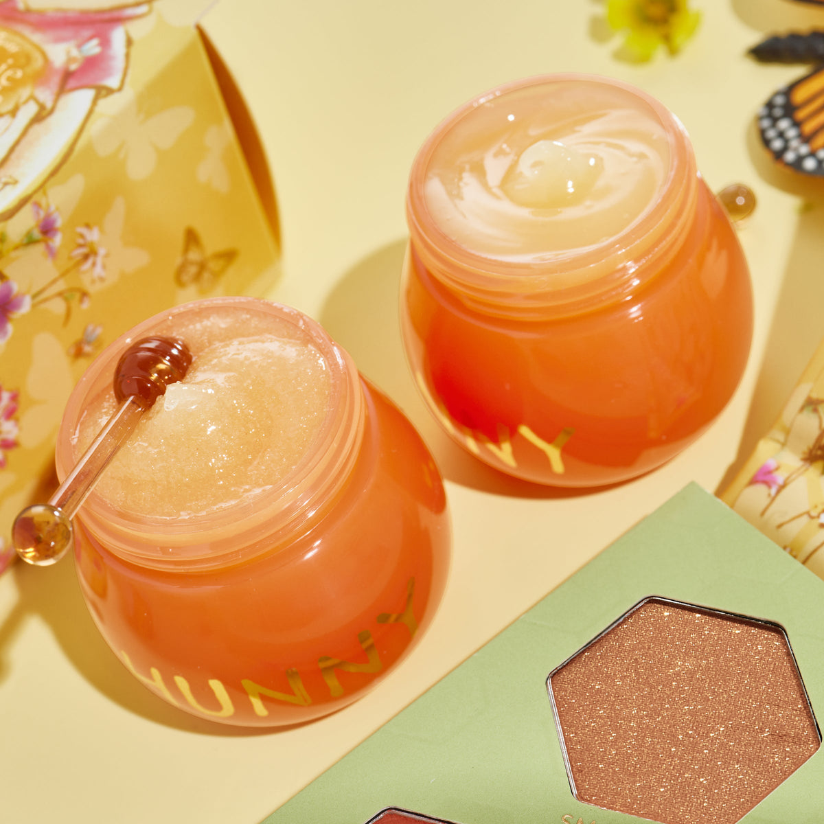 Sweet as Hunny Eye & Lip Set honey-pot orange jars sit open, revealing a sugar lip scrub and glossy balm with a mini honey dipper, beside a green shimmery eyeshadow palette and floral butterfly packaging on a pastel yellow surface; ColourPop Lip Care Kit.