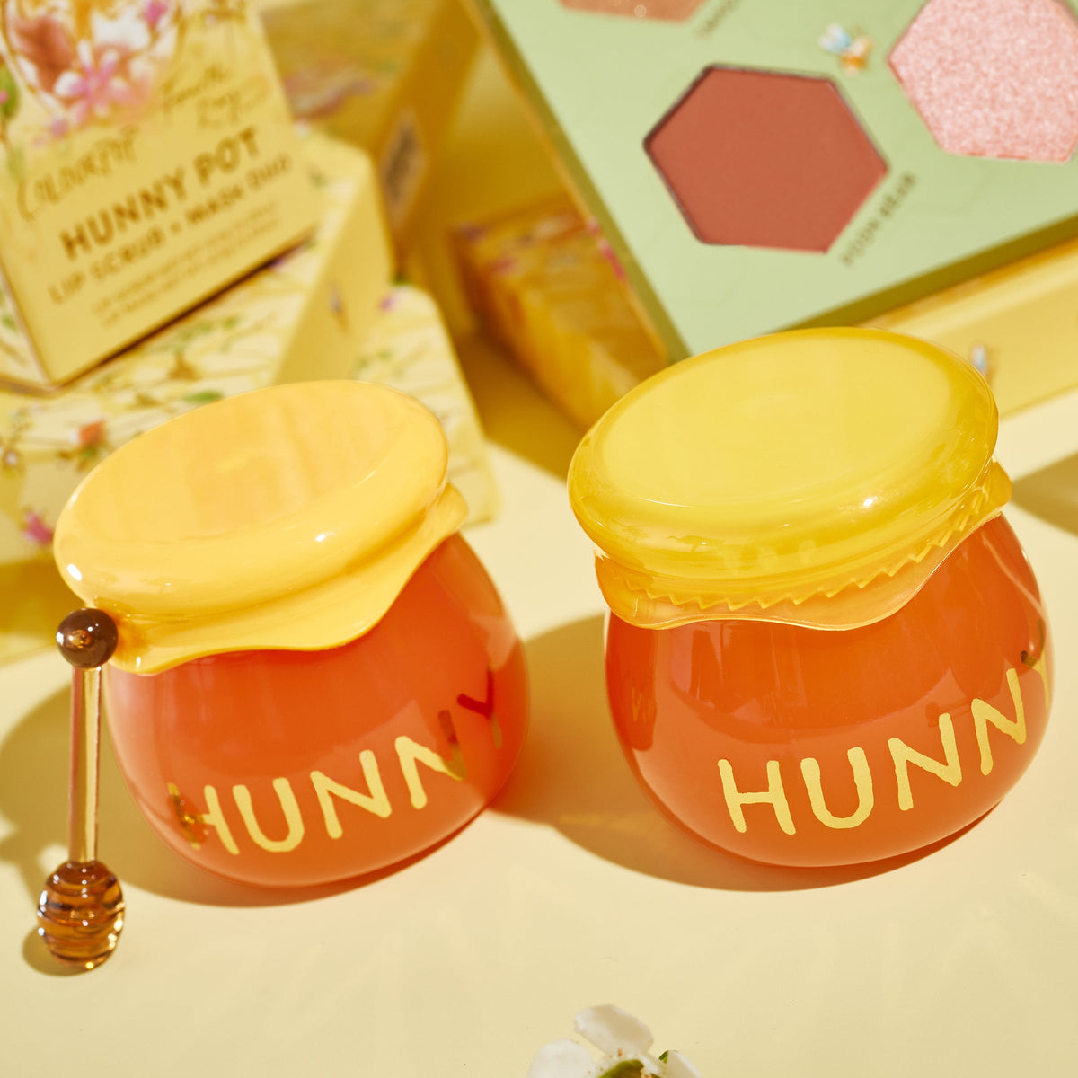 Hunny Pot lip care kit by ColourPop - two honey jar–shaped pots with yellow lids and HUNNY lettering - sit on a pale yellow surface beside a mini honey dipper, floral box, and makeup palette, showcasing the lip scrub and mask duo.