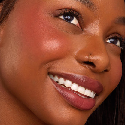 Canon Dr Ultra Blotted Lip on a smiling model, lips softly blurred in a deep rosy mauve finish, showcasing a lightweight stain, with cheeks glowing from a complementary flush of ColourPop Cream Blush in a crisp studio close-up.