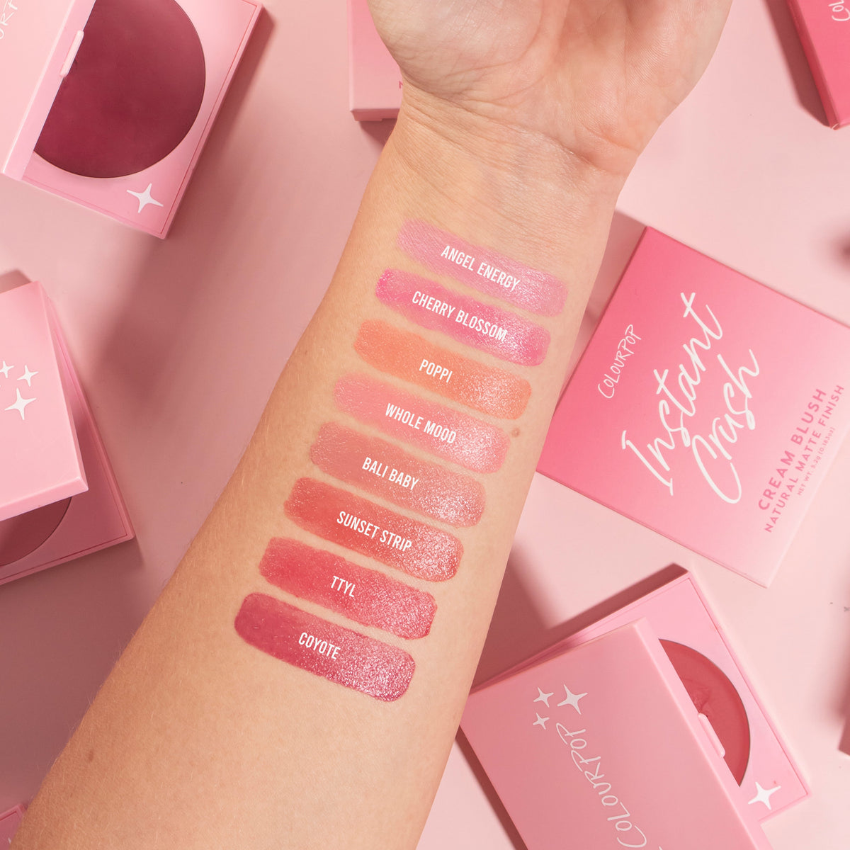 Forearm displays eight ColourPop Cream Blush swatches from the Instant Crash line - Angel Energy, Cherry Blossom, Poppi, Whole Mood, Bali Baby, Sunset Strip, TTYL, and Coyote - shown on light skin with pink packaging compacts around.