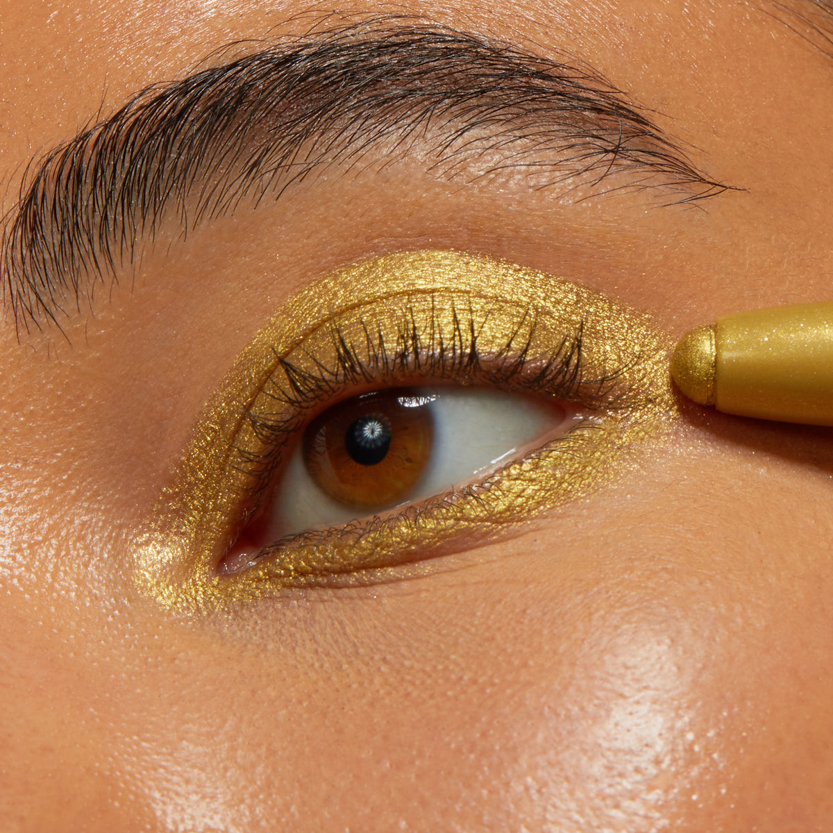 ColourPop Flex Shadow Stix applies a rich metallic gold shade on a brown eye, tracing the upper and lower lash lines and covering the lid for a bold, shimmery finish in a tight beauty close-up.