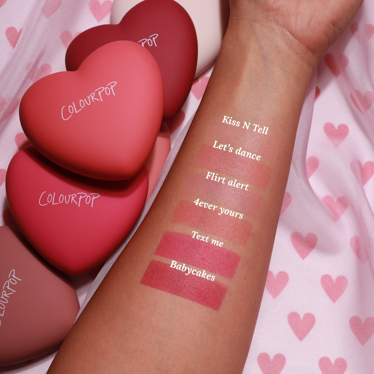 ColourPop Kiss 'N Tell pressed powder blush swatched on a forearm, showing a soft rosy hue beside labeled shade swatches, with matte heart-shaped ColourPop compacts in pink and red resting on a pink heart-print fabric.